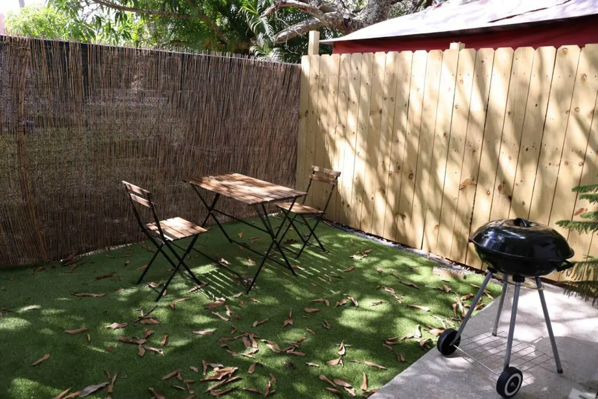 This image captures a private outdoor patio featuring a wooden table with two folding chairs, a BBQ grill, bamboo fencing for privacy, and artificial grass flooring. The scene is styled for relaxed outdoor living and dining, with subtle touches that evoke a tranquil, sun-filled Miami retreat.