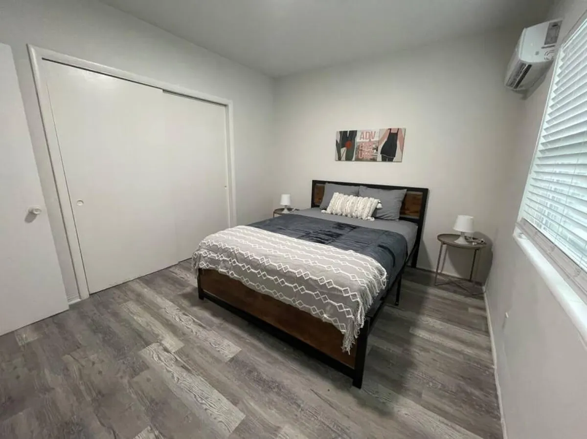 The image shows a bright and airy bedroom with a plush queen bed adorned with a stylish patterned throw and pillows, flanked by two bedside tables with modern lamps. Natural light pours in through a window with blinds, and a sleek air conditioning unit ensures year-round comfort. The overall space feels calm, clean, and inviting—perfect for relaxation after a day in Miami.