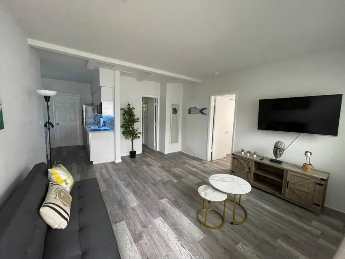 A bright, contemporary living area featuring a gray sofa with decorative pillows, a mounted flat-screen smart TV, and a set of marble-topped nesting coffee tables. The space is accented by a tall potted green plant near the kitchen, with natural light enhancing the calm, comfortable ambiance. The kitchen area is visible, suggesting a seamless, open studio layout.