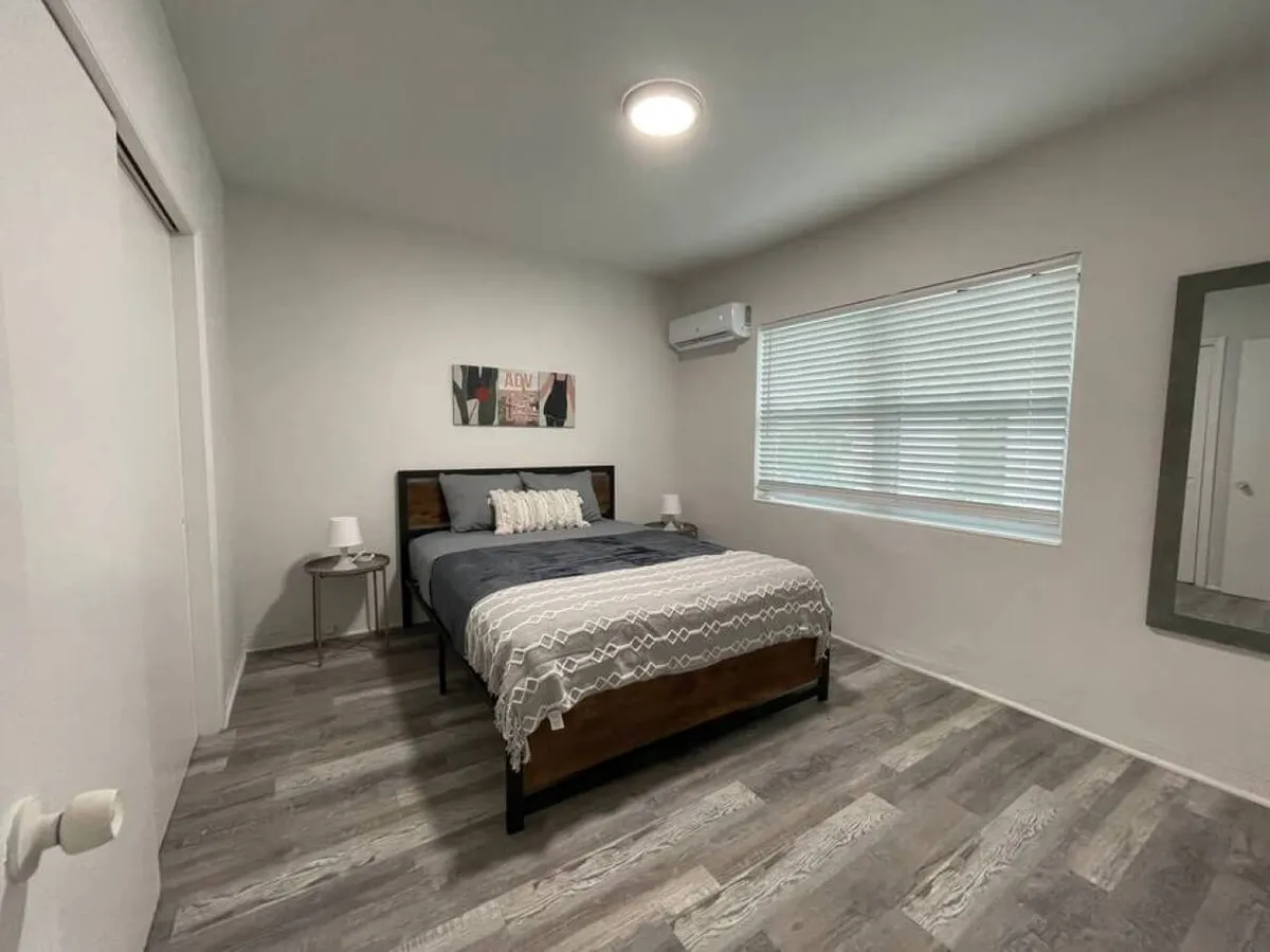 A restful sleeping area with a queen bed dressed in textured bedding, minimalist bedside tables with lamps, and a large window with blinds that lets in abundant daylight. The space is finished with modern decor, including framed artwork above the bed, creating a tranquil retreat for rest and relaxation.
