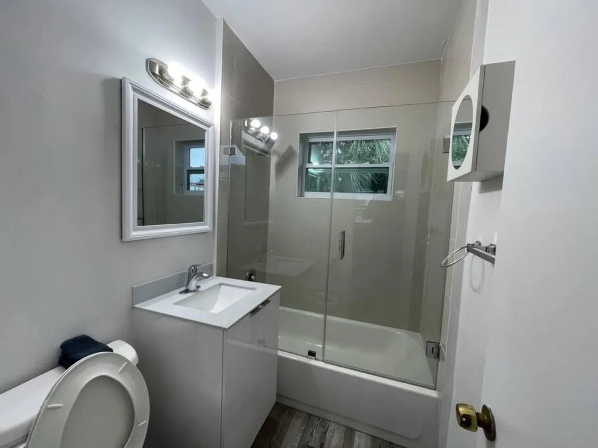 The photo captures a bright, fresh bathroom interior featuring a white vanity with a sink, a glass-enclosed bathtub/shower, and a large mirror reflecting natural light from a small window with greenery visible outdoors. The space is clean, modern, and exudes a sense of comfort and functionality.