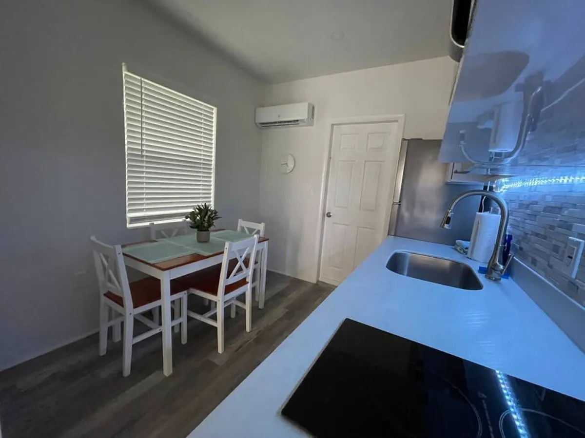 This image displays a bright and inviting modern kitchen with an integrated sink and stove, a small dining table set for four with a potted plant centerpiece, and a window allowing gentle daylight to fill the space. The kitchen is neatly organized, showcasing modern appliances and a cozy dining area adjacent to the living space. The presence of a wall-mounted air conditioning unit suggests year-round comfort, while the proximity to a private patio hints at easy transitions for indoor-outdoor dining.